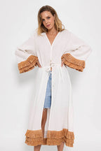Woman wearing white long cotton kimono with tan raffia fringe details on sleeves and hem over denim shorts