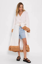 Woman wearing long white kimono with tan fringe detailing paired with denim shorts and black sandals