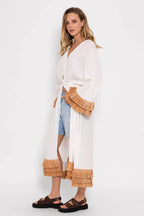 Woman wearing a long white kimono with raffia fringe details, paired with denim shorts and brown sandals.