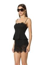 Woman wearing a black lace peplum top and skirt set with spaghetti straps and large sunglasses in a studio.