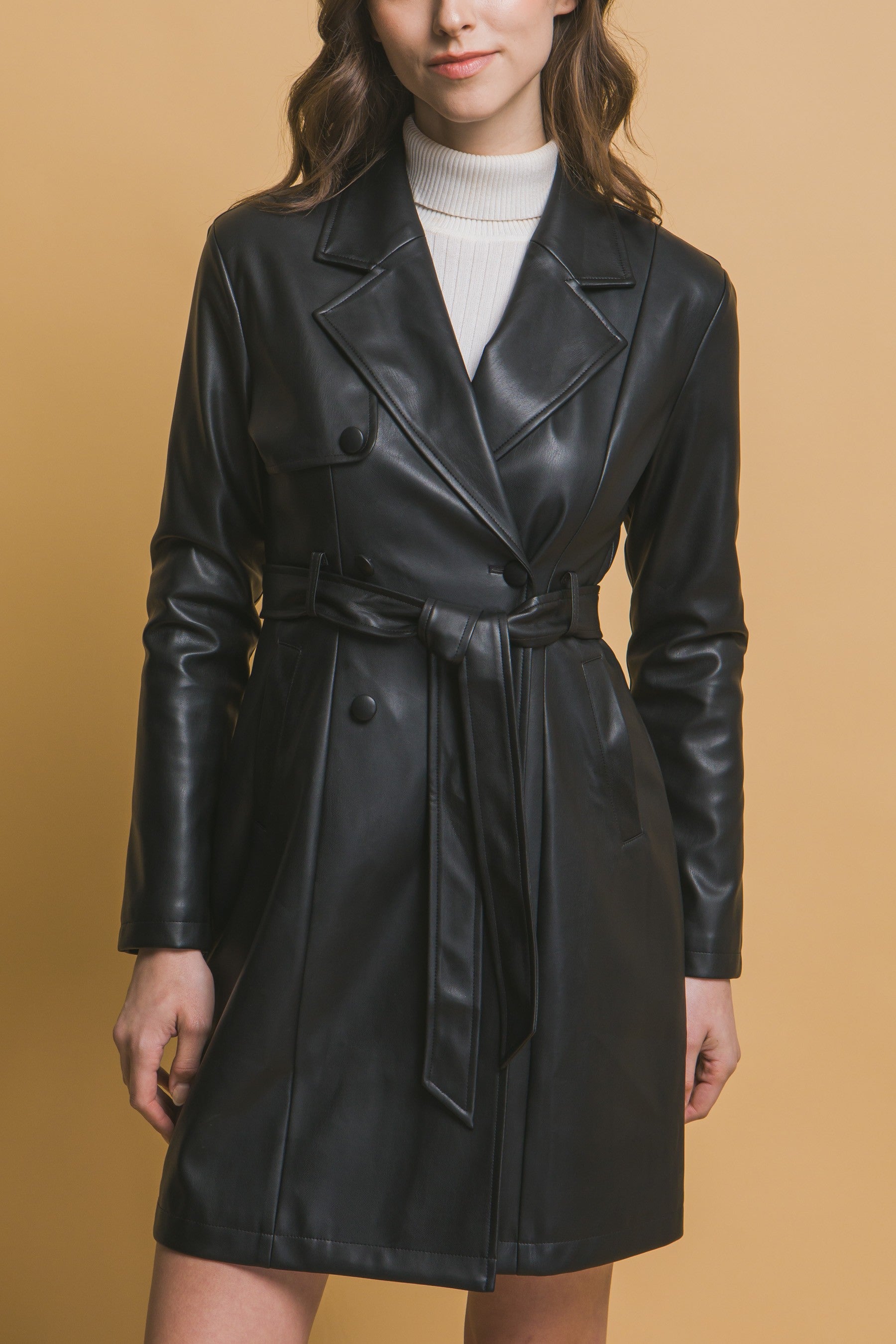 Woman wearing a black vegan leather belted jacket over a white turtleneck against a beige background
