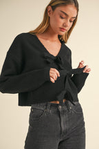 Woman wearing a black knit sweater top with bow ties over dark high-waisted jeans, adjusting the front bow detail.