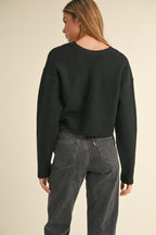 Woman wearing a black knit sweater with back bow detail paired with high-waisted dark gray jeans, shown from behind.