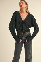 Woman wearing black knit sweater with bow ties down the front paired with dark high-waisted jeans in a studio setting.