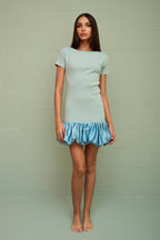 Woman wearing a fitted ribbed mini dress with a voluminous satin bubble hem in light blue tones against a plain wall.