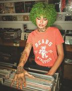 Woman with green curly hair wearing a red Bears Athletics tee browsing vinyl records in a vintage store.