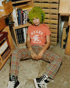 Woman with green hair wearing a Bears Athletics orange tee and plaid pants sitting on a workshop floor in black sneakers