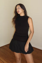 Woman wearing a sleeveless black ribbed top paired with a black bubble mini skirt in a simple indoor setting.