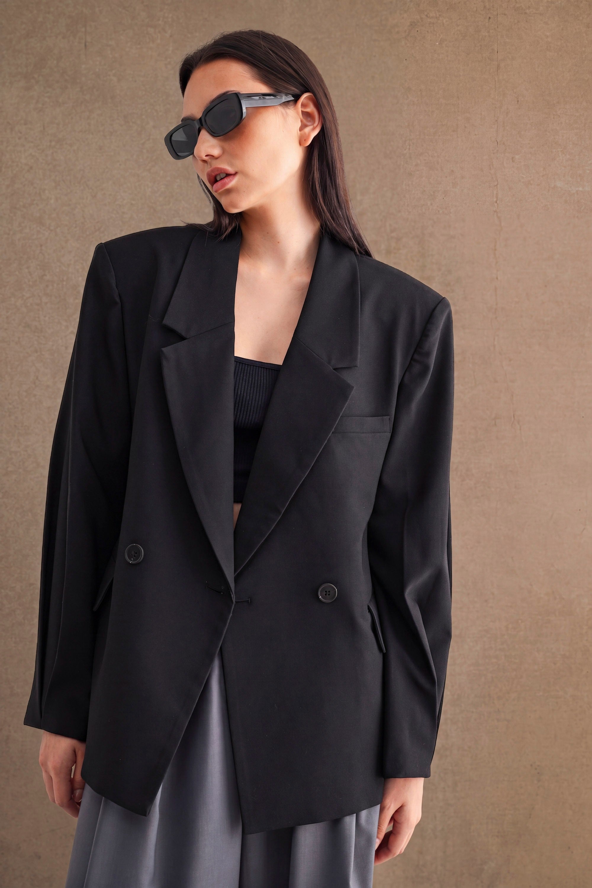 Woman wearing oversized black blazer and sunglasses styled with a black top and gray skirt in minimalist setting