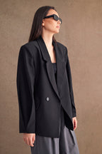 Woman wearing an oversized black blazer with structured shoulders and sunglasses against a plain background.