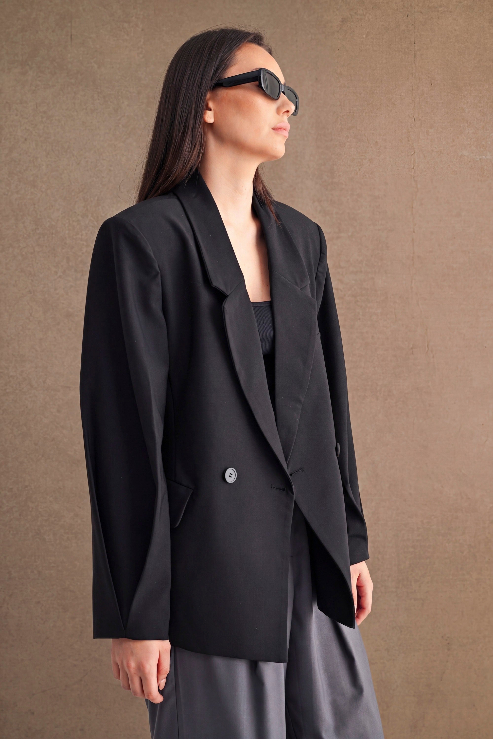 Woman wearing an oversized black blazer with structured shoulders and sunglasses against a plain background.