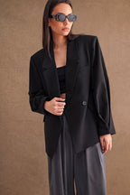 Woman wearing an oversized black blazer and sunglasses paired with loose gray pants against a brown background