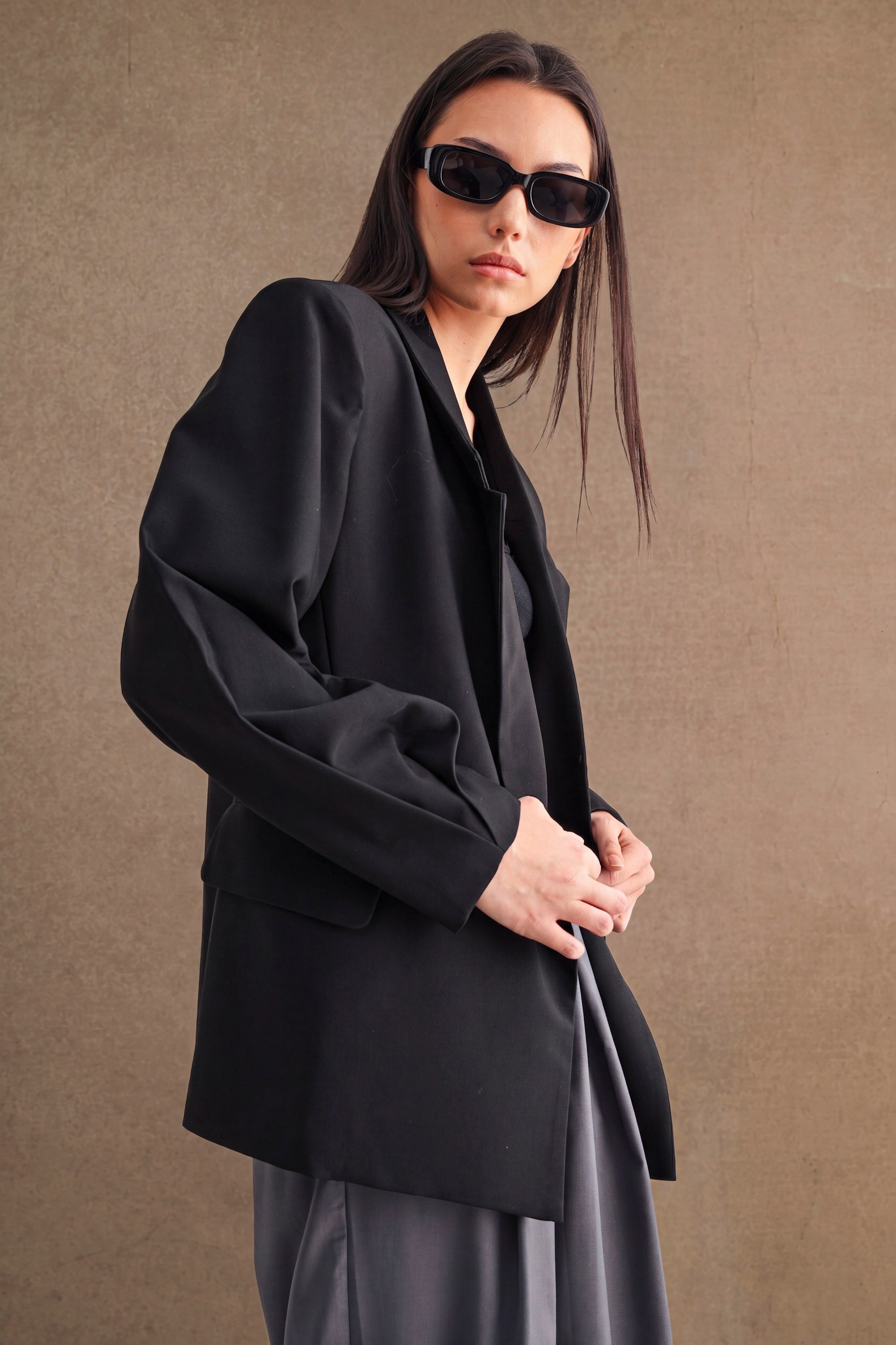 Woman wearing oversized black blazer and sunglasses standing against a plain brown background in women's clothing style.