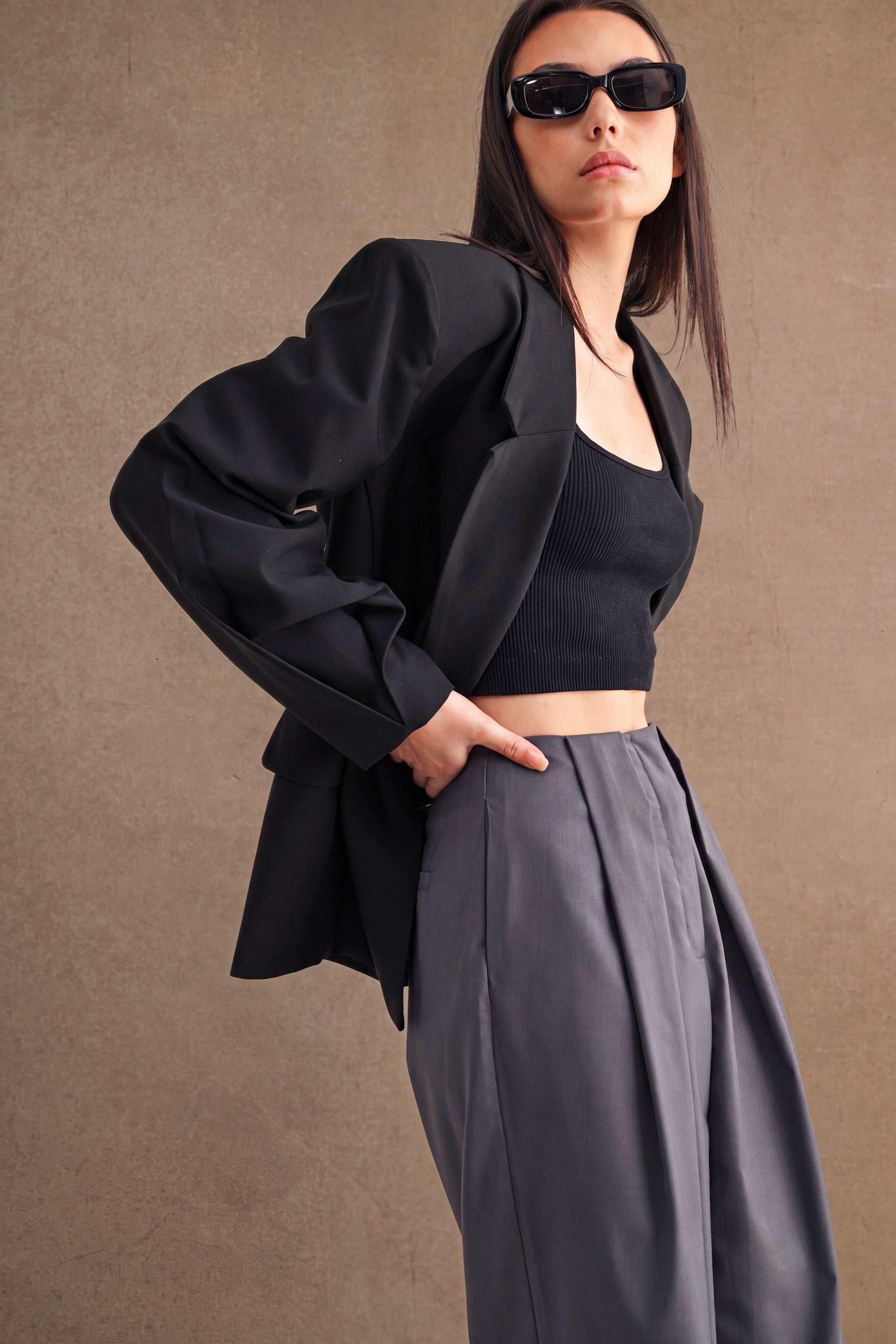 Woman wearing an oversized black blazer over a black crop top with high-waisted gray pants in a studio setting