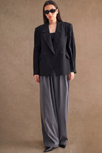 Woman wearing an oversized black blazer and wide-leg gray pants with sunglasses against a plain background
