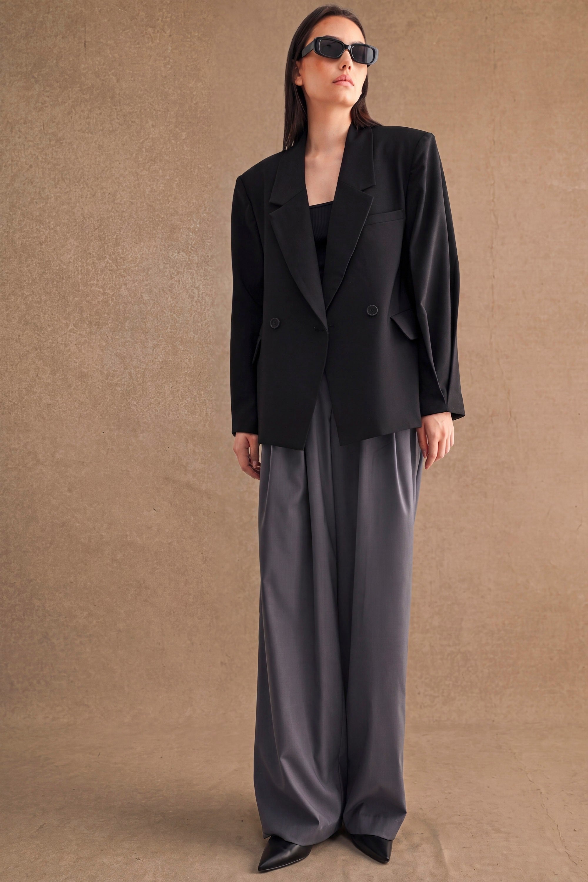Woman wearing oversized black blazer paired with wide-leg gray pants and black sunglasses against neutral background
