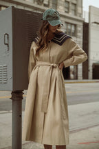 Woman wearing a beige belted trench coat with navy sailor collar and a green baseball cap on city street