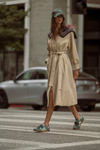 Woman wearing beige trench coat with belt and green cap walking across city street wearing sneakers
