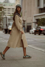 Woman wearing beige trench coat with dark collar detail and green sneakers walking on city street
