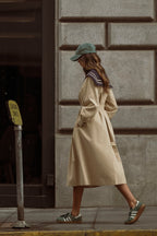 Woman walking in beige trench coat paired with green cap and casual sneakers on city street