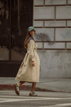 Woman wearing beige trench coat with navy collar and green cap crossing a city street in stylish women's clothing