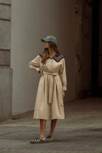 Woman wearing a beige trench coat with a navy collar detail, paired with a green cap and casual sneakers outdoors