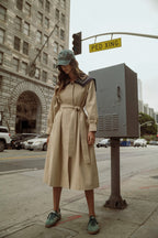 Woman wearing a beige belted trench dress with a navy collar detail and green sneakers on a city sidewalk