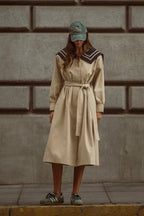 Woman wearing beige belted trench coat with dark collar detail and green cap standing against stone wall