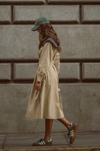 Woman wearing a beige trench coat with a belt, green cap, and casual sneakers walking along a stone wall background.