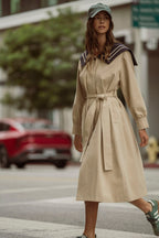 Woman wearing beige belted trench dress walking across city street with baseball cap and sneakers