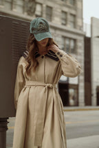 Woman wearing a beige trench coat with a belt and a green cap, standing on a city street in casual women's clothing.