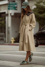 Woman wearing beige belted trench coat and green cap walking on city street in casual style women's clothing