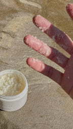 Close-up of fingers with grainy natural body scrub texture next to an open jar on a stone surface