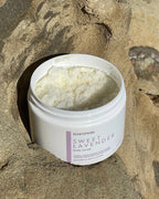 Open jar of natural lavender-scented body scrub placed on textured rock surface in sunlight
