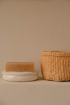 Natural handmade soap bar on white ceramic dish next to woven straw basket on beige surface