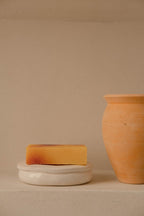 Natural handmade soap bar on white ceramic dish next to a terracotta vase on beige background