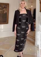 Woman wearing black strapless midi dress with sequin bow patterns and a black blazer draped over shoulders in elegant room