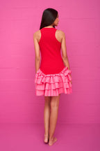 Woman wearing a sleeveless red mini dress with layered pink ruffles, standing barefoot against a pink wall background