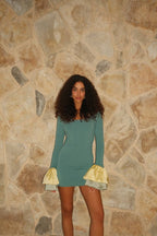 Woman wearing a green mini dress with long sleeves and yellow satin ruffle cuffs standing against a stone wall
