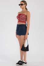 Woman wearing a striped red crop tube top with denim mini skirt, black handbag, black sunglasses, and black sneakers.