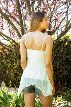Woman wearing a white boho tank top with spaghetti straps and denim shorts standing outdoors in a garden setting