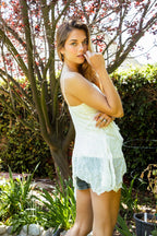 Woman by garden tree wearing a white boho-style tank top and denim shorts in natural sunlight