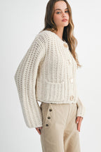 Woman wearing a cream chunky knit sweater with gold buttons and high-waisted beige pants in a studio setting