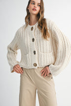 Woman wearing a cream chunky knit sweater with large gold buttons and beige pants in a fashion pose