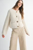 Woman wearing a chunky knit cream sweater with large gold buttons and beige high-waisted pants.