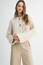 Woman wearing a chunky cream knit sweater with large gold buttons paired with beige high-waisted pants.
