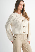 Woman wearing chunky cream knit sweater with large gold buttons and beige pants with button details