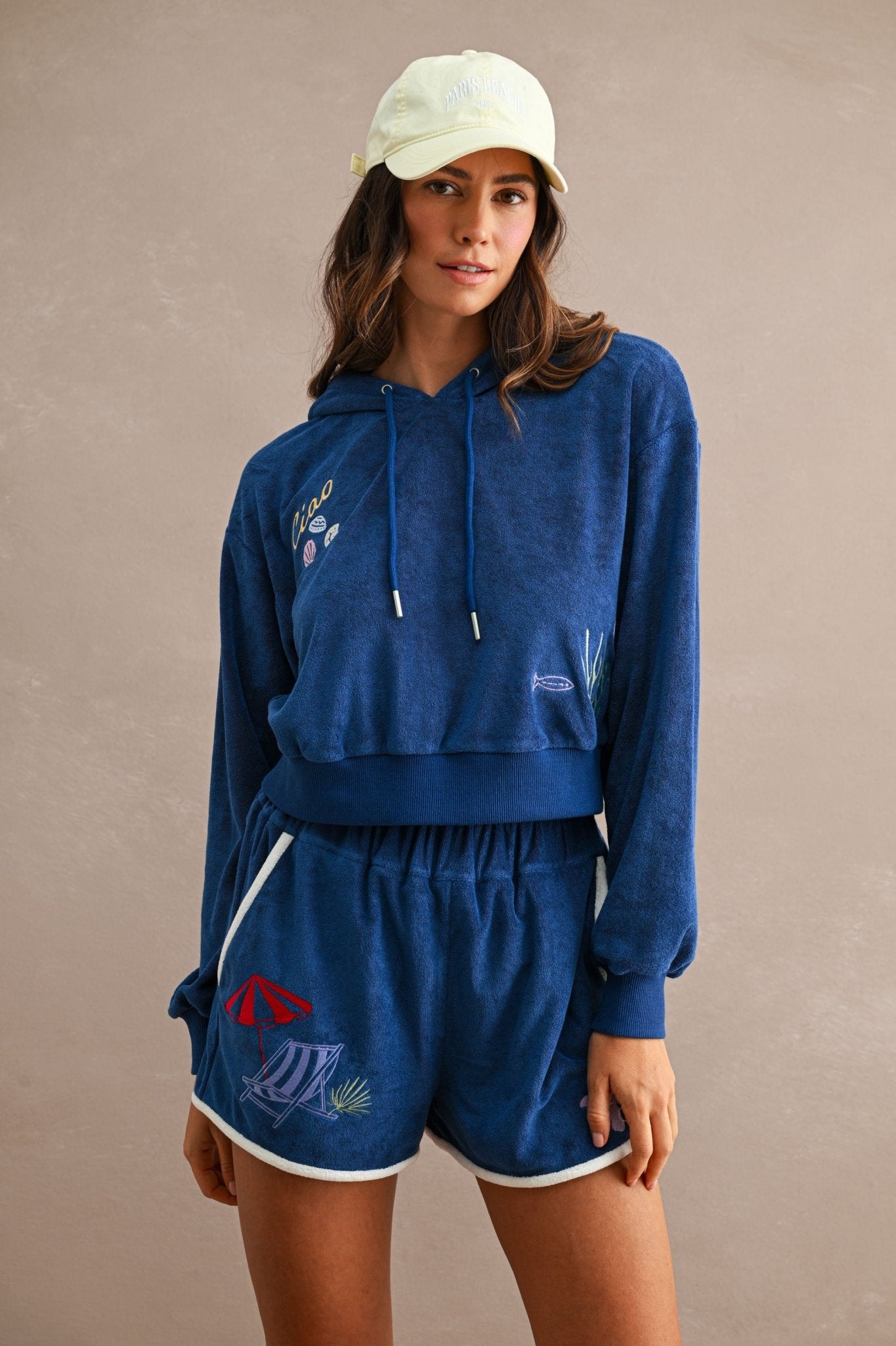Woman wearing a navy blue Ciao Terry Hoodie with a drawstring hood and matching shorts featuring colorful embroidered beach designs, paired with a light beige cap.