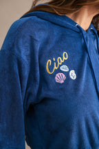 Close-up of a blue terry cloth hoodie with embroidered "Ciao" and colorful beach-inspired shell designs on the chest.