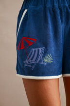 Close-up of blue terry cloth shorts with embroidered beach chair, umbrella, and palm leaf detail on women's clothing.